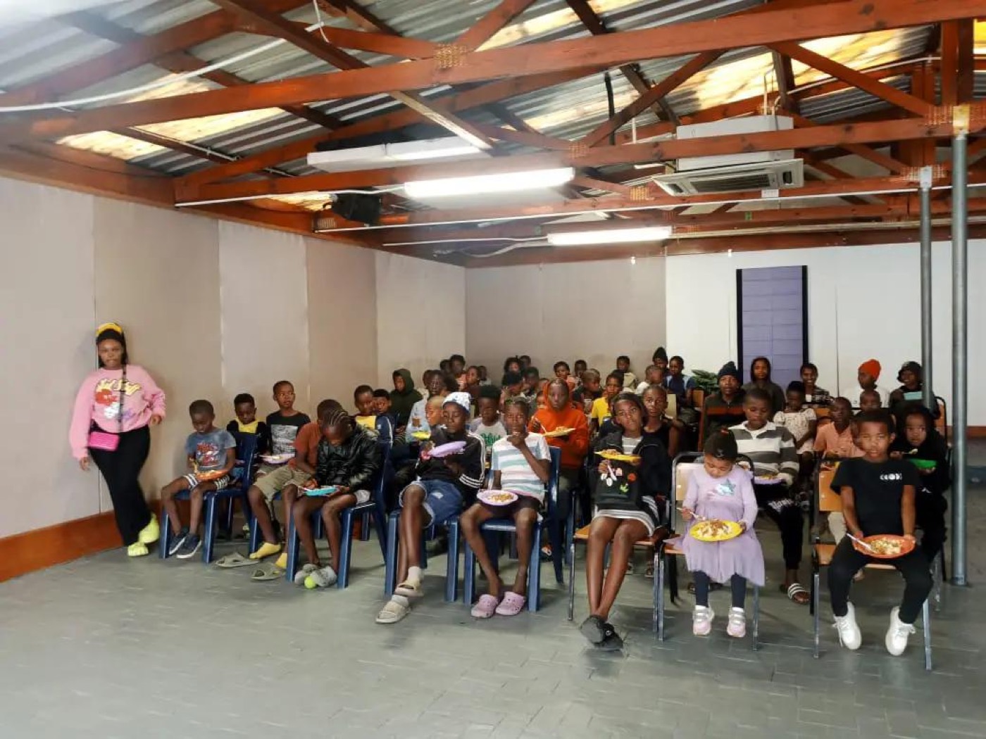 A full hall of children seated with plates on their laps